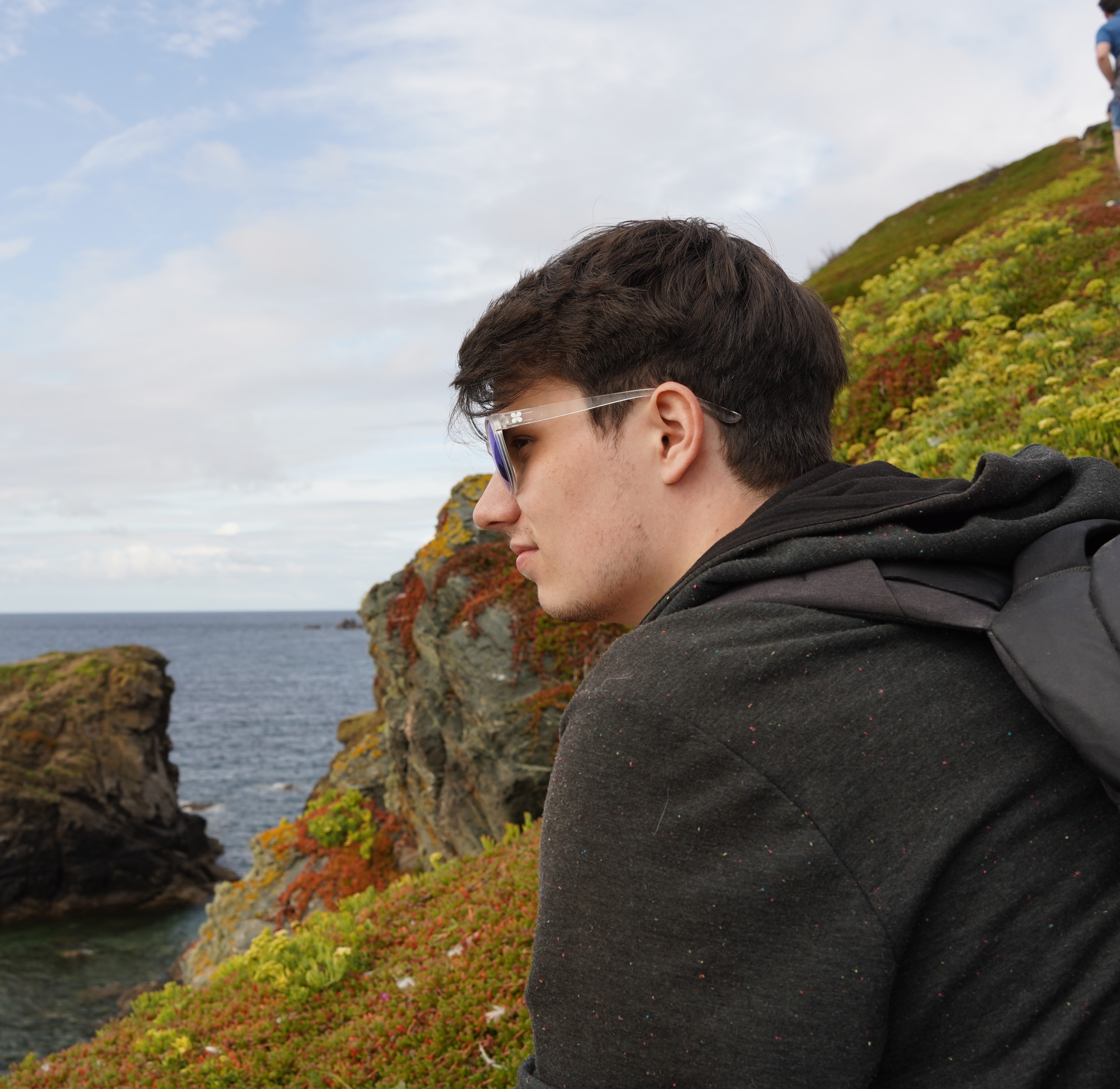 A photo of myself sat on the green cliffs of Cornwall in the UK with the ocean & sky in the background.